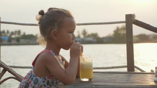 Funny little girl is sitting in a riverside cafe at a table and drinking juice — Stock Video