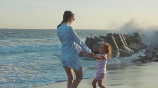Мать крутит свою маленькую дочь на песчаном пляже в теплых лучах заката . — стоковое видео