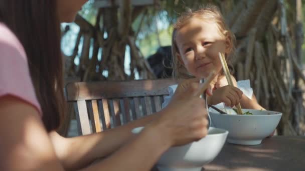 Moeder en dochter eet noodles soep via stokjes in de open lucht strand café — Stockvideo