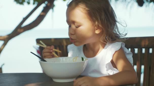 Gyermek lány eszik tészta leves keresztül pálcika, a szabadtéri strand kávézó — Stock videók