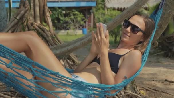 Молодая женщина лежит на гамаке со смартфоном на песчаном пляже в замедленной съемке — стоковое видео