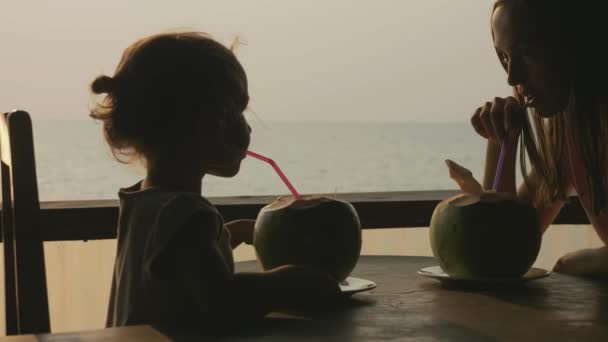 Young mother and daughter sits at seaview cafe and drinks coconats together — Stock Video