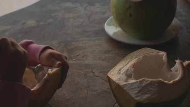 Маленька мила дівчинка їсть кокос ложкою в кафе з видом на море в повільному русі — стокове відео