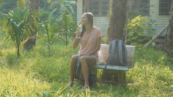 En ung kvinne sitter på benken i landsbygda og drikker vann. – stockvideo