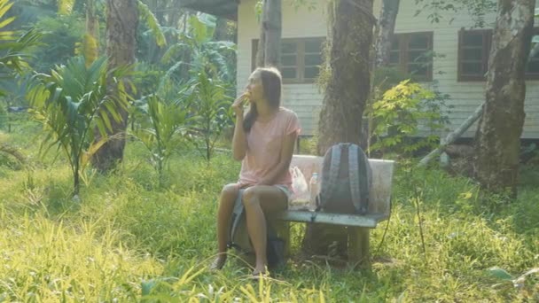 Mujer joven sentada en el banco en el pueblo rural asiático y comiendo plátano — Vídeos de Stock