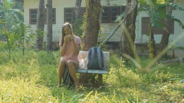 En ung kvinne sitter på benken i en landlig landsby og spiser mat. – stockvideo
