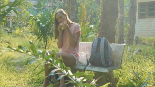 Jeune femme assise sur le banc à la campagne asiatique village et manger collation — Video