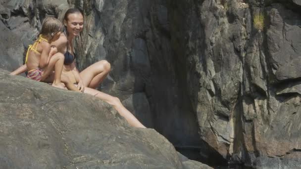 Молодая мама с милой маленькой дочкой отдыхает на скальном берегу горной реки — стоковое видео
