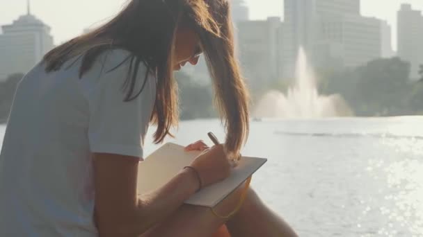 Mujer joven sentada en el banco en el parque y escribiendo en el diario, primer plano — Vídeo de stock