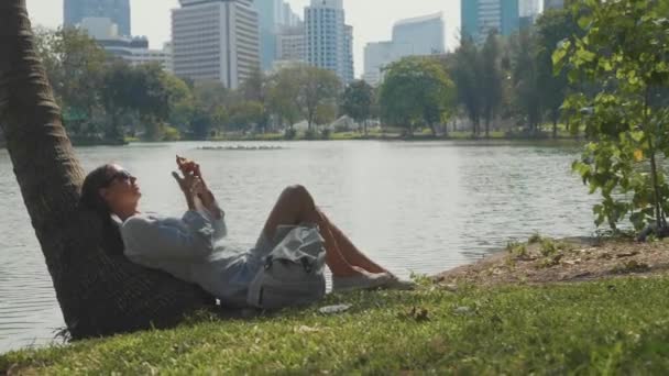 Junge Frau liegt mit Handy unter Palme im Park — Stockvideo