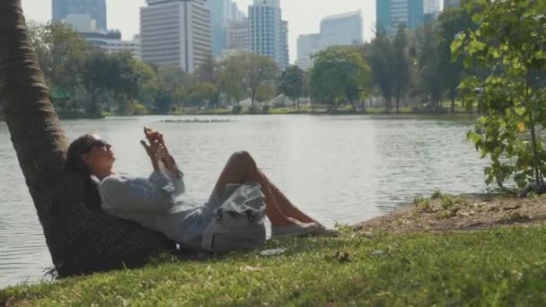 Mujer joven acostada bajo la palmera usando teléfono móvil en un parque — Vídeo de stock