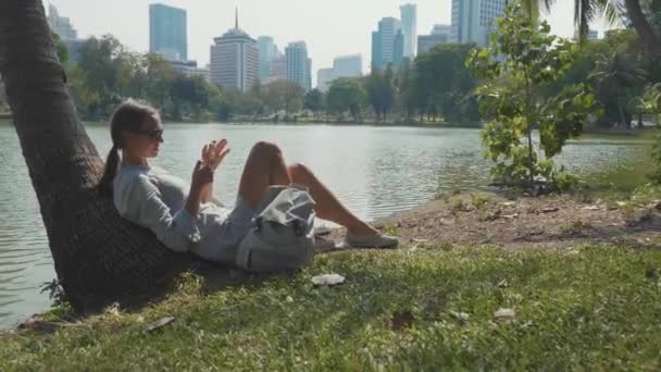 Mujer joven acostada bajo la palmera usando teléfono móvil en un parque — Vídeo de stock