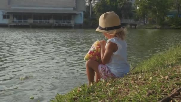 Little girl in country style hat feeding fishes in a park lake in slow motion — Stock Video