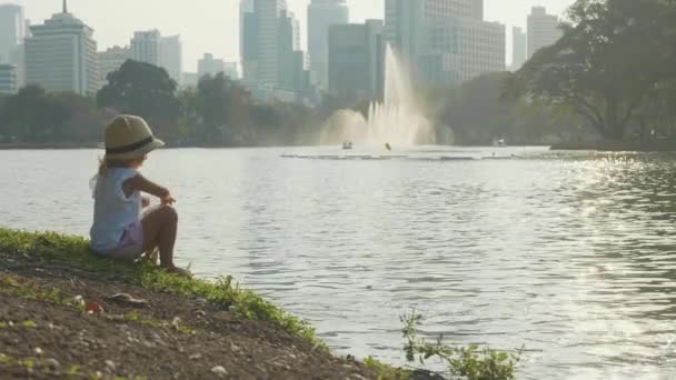 Klein meisje in land stijl hoed voeden van vissen in een park lake in slow motion — Stockvideo