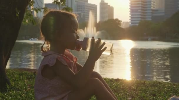 Kind trinkt Wasser im Park mit See und Wolkenkratzern im Hintergrund — Stockvideo