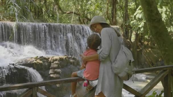 En ung mamma och hennes söta dotter vilar mot kaskad av ett vattenfall — Stockvideo