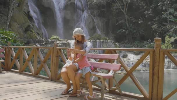 Uma jovem mãe e sua linda filha descansando contra a cascata de uma cachoeira — Vídeo de Stock