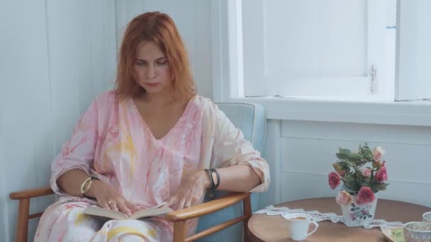 Jeune femme charmante assise sur la chaise et le livre de lecture à côté de la fenêtre à la maison — Video