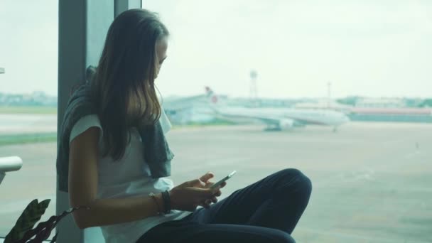 Молодая женщина использует смартфон в аэропорту с самолетом на заднем плане — стоковое видео