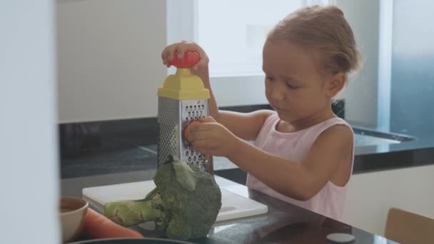 Χαριτωμένο μικρό παιδί κορίτσι τρίβει το καρότο σε ένα τρίφτη στην εγχώρια κουζίνα. — Αρχείο Βίντεο
