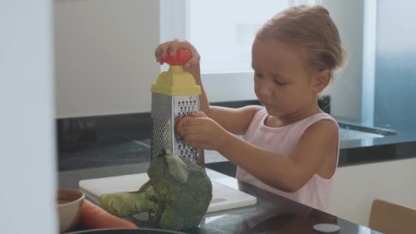 Linda niña frotando zanahoria en un rallador en la cocina doméstica . — Vídeo de stock