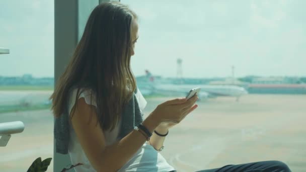 Wanita muda menggunakan telepon pintar di bandara dengan pesawat terbang di latar belakang — Stok Video