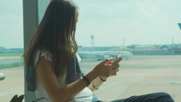 Jonge vrouw is met behulp van slimme telefoon op luchthaven met vliegtuig op de achtergrond — Stockvideo