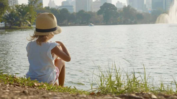 Menina em estilo country chapéu sentado na margem do lago no parque da cidade — Fotografia de Stock
