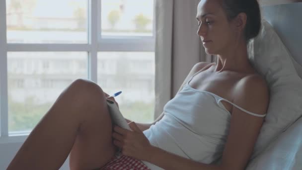 Молодая женщина лежит на кровати и пишет утренние страницы в дневнике, замедленной съемки — стоковое видео