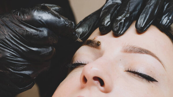 The master in black gloves is adding some drops of the brown pigment on the clients eyebrow after the microblading procedure to make a mask above the brow. Close-up.