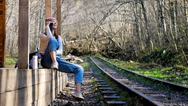 Mulher mochileiro jovem está sentado na estação ferroviária e falando no smartphone — Fotografia de Stock