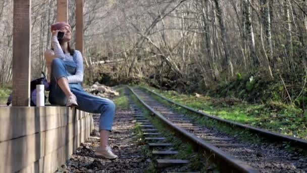 Mujer joven mochilero está sentado en la estación de tren y hablando en el teléfono inteligente — Vídeo de stock