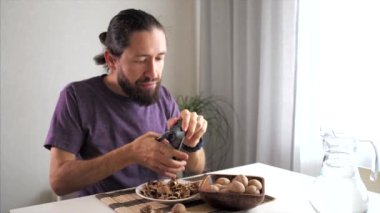 Genç sakallı adam evde ceviz kırıyor ve yiyor. Evde masada otururken fındık kırmak için çelik fındık kırıcı kullanıyor..