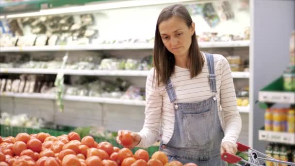 Молода жінка в джинсовому загальному вибирає свіжі помідори в продуктовому магазині . — стокове відео