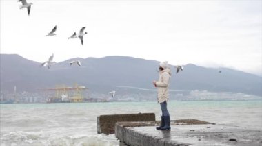 Young woman is feeding a seagulls. Seashore in storm windy weather
