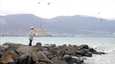Young woman is feeding a seagulls. Seashore in storm windy weather