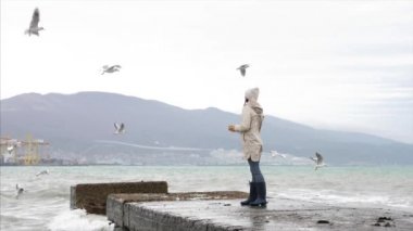 Young woman is feeding a seagulls. Seashore in storm windy weather