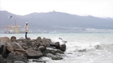 Woman is walking by empty seafront in windy weather with crashing sea waves
