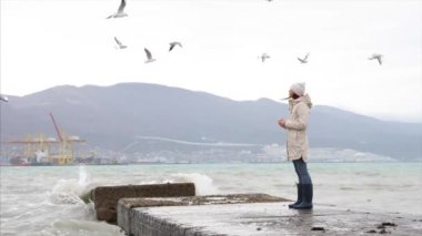Young woman is feeding a seagulls. Seashore in storm windy weather