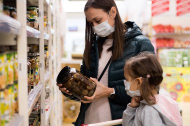 Tıbbi maskeli kız çocuğu olan genç bir kadın süpermarketten konserve yiyecek alıyor.