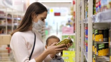 Tıbbi maskeli kız çocuğu olan genç bir kadın süpermarketten konserve yiyecek alıyor.
