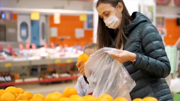 Mladá žena s dítětem dívka v lékařských maskách kupuje pomeranče v supermarketu — Stock video