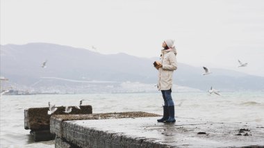 Young woman is feeding a seagulls. Seashore in storm windy weather