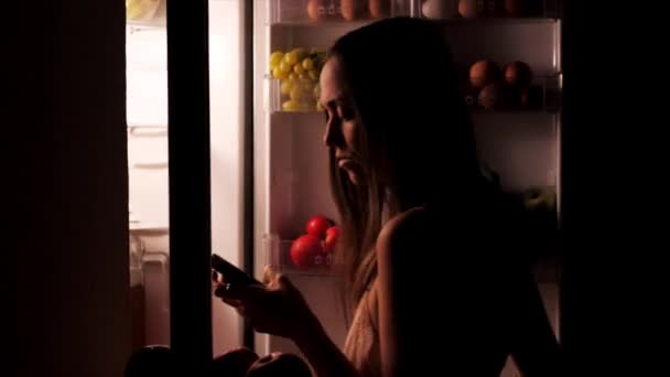 Женщина, использующая смартфон и перекусывающая ночью — стоковое видео