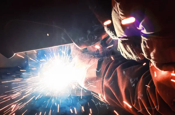 Lasser in een beschermend masker in een donkere werkvloer weld metalen onderdelen. Door het lassen van vonken vliegen in verschillende richtingen — Stockfoto