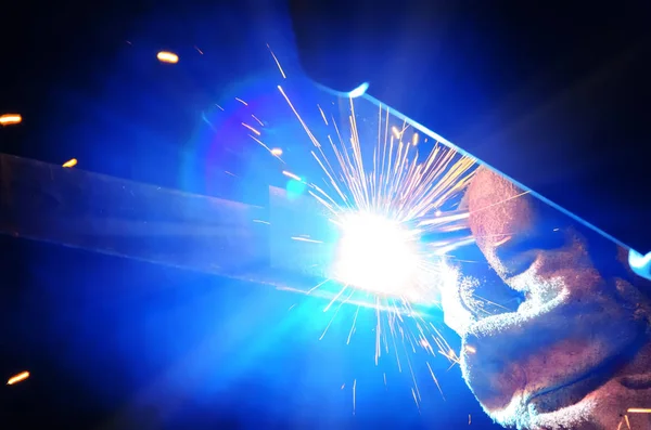 Lasser in een beschermend masker in een donkere werkvloer weld metalen onderdelen. Door het lassen van vonken vliegen in verschillende richtingen — Stockfoto