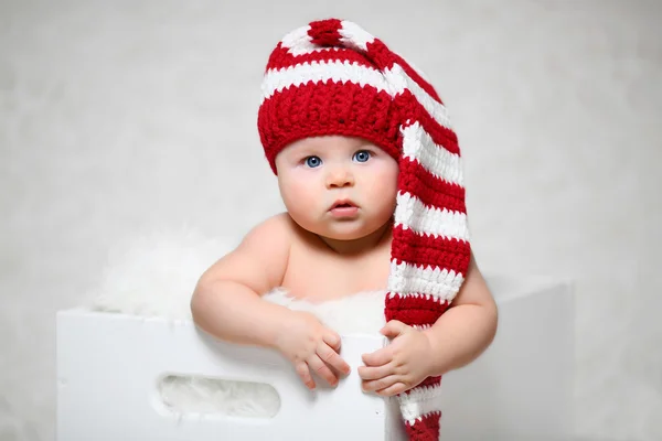 Um bebê de Natal sentado em uma caixa de madeira branca vestindo um chapéu de gnomo listrado vermelho e branco — Fotografia de Stock