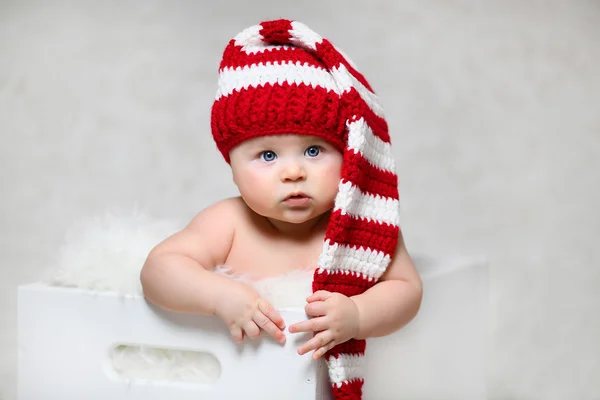 Um bebê de Natal sentado em uma caixa de madeira branca vestindo um chapéu de gnomo listrado vermelho e branco — Fotografia de Stock