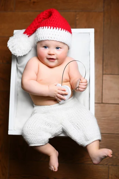 Um bebê de Natal deitado em uma caixa branca fofa usando um chapéu de cauda vermelha e calças brancas e segurando fones de ouvido — Fotografia de Stock