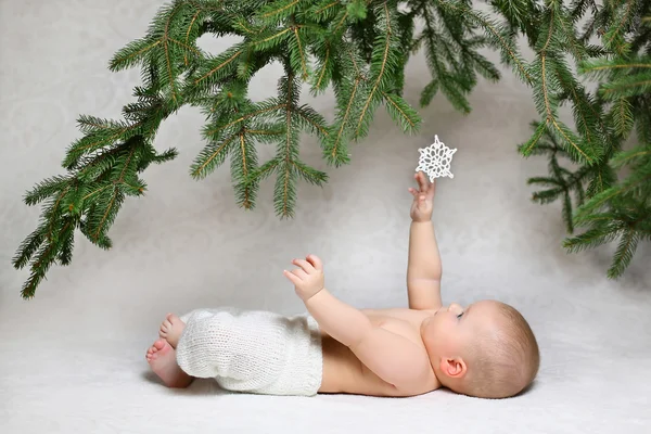 Bebê bonito deitado unger um ramo de árvore de natal em calças brancas de malha tentando chegar a um floco de neve — Fotografia de Stock